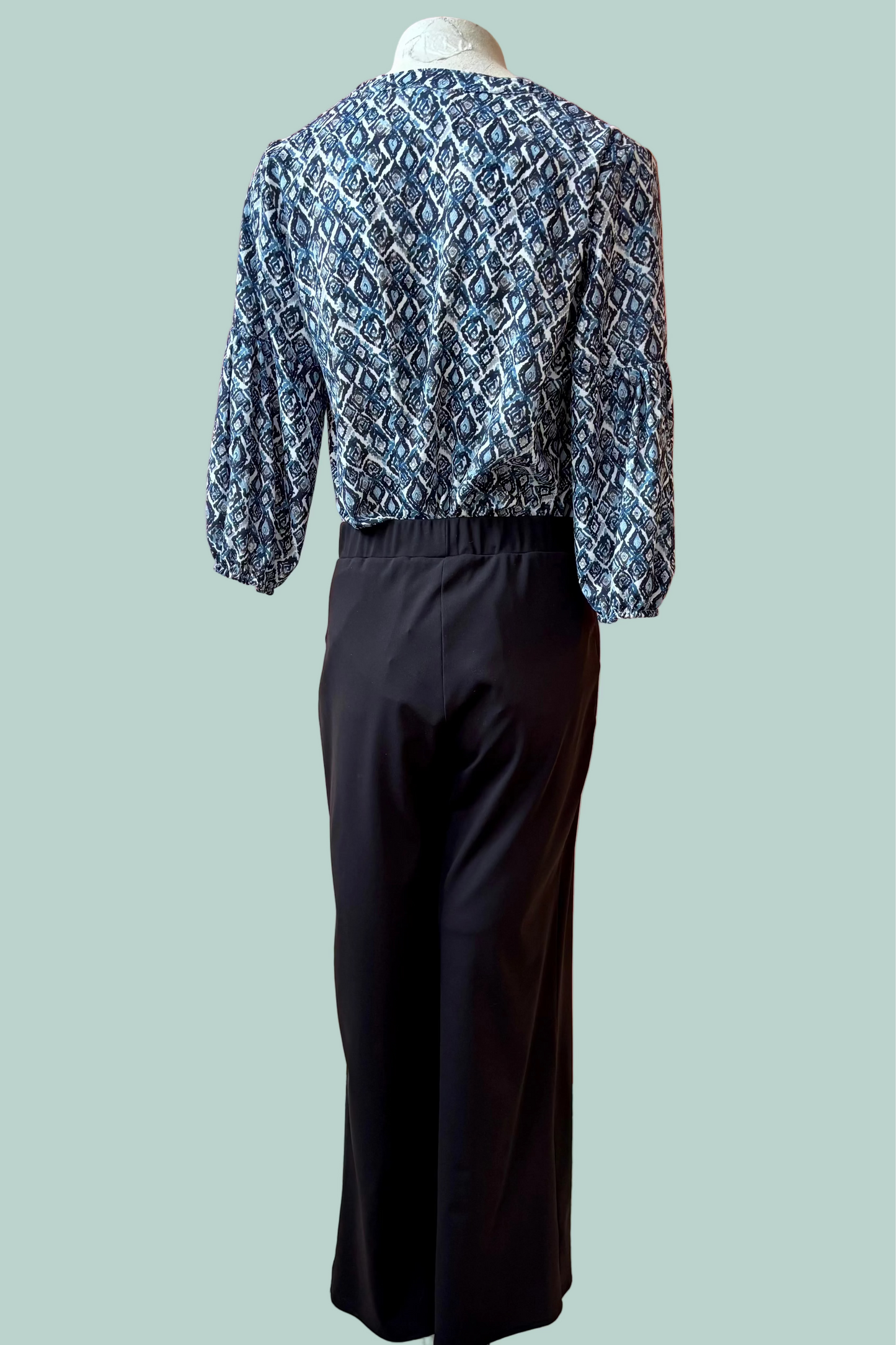 Back view of a mannequin wearing a the Gloria Pants by Pure Essence in Black with a blue patterned top on a light teal 