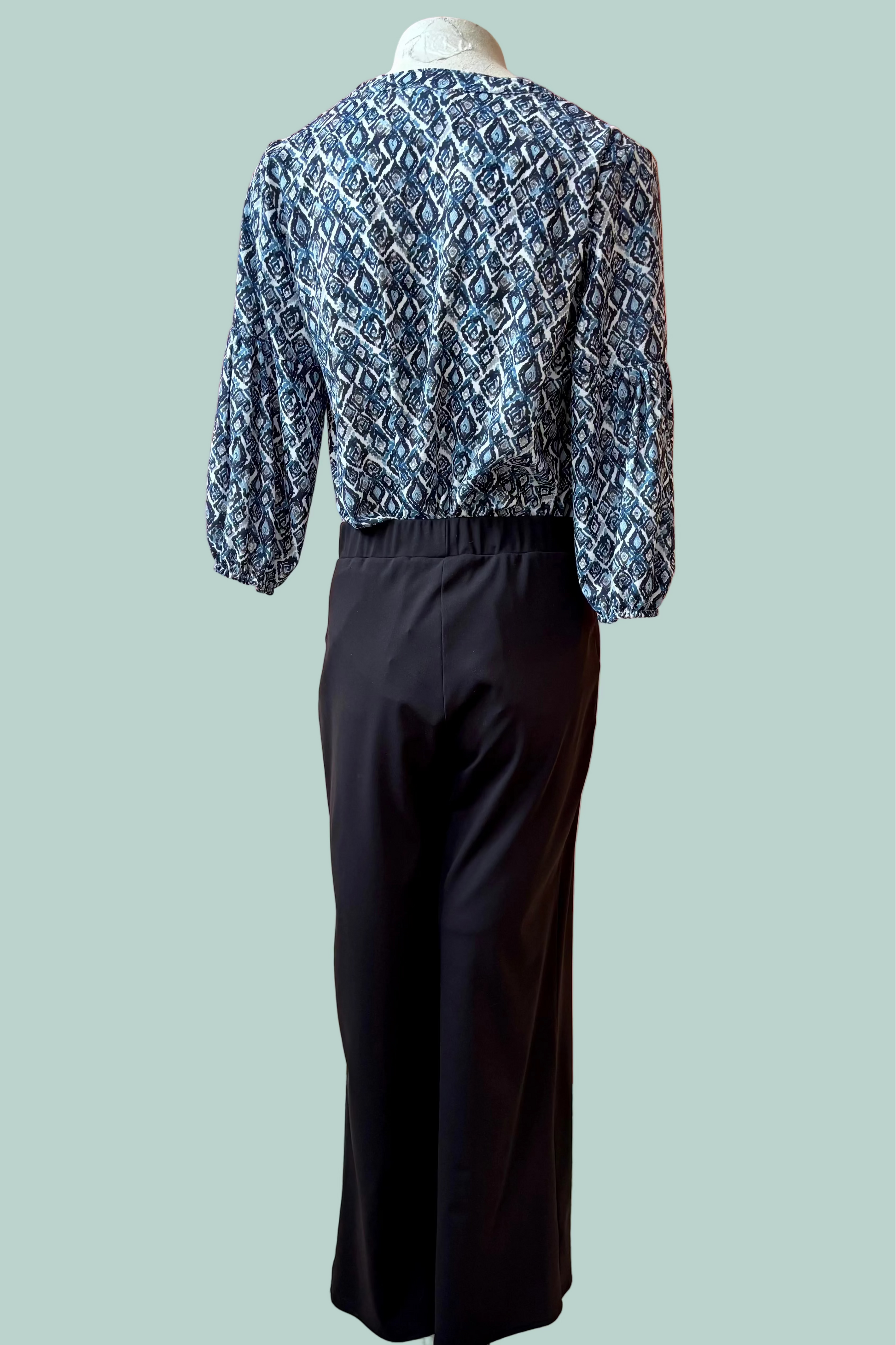 Back view of a mannequin wearing a the Gloria Pants by Pure Essence in Black with a blue patterned top on a light teal 