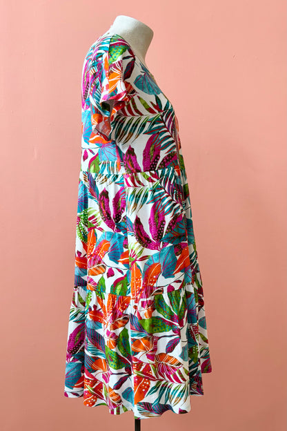 Side view of the Grayson Dress by Pure Essence in Peacock Palms, a knee-length baby doll style dress with a tiered skirt and short sleeves, is shown on a mannequin against a coral  wall. 