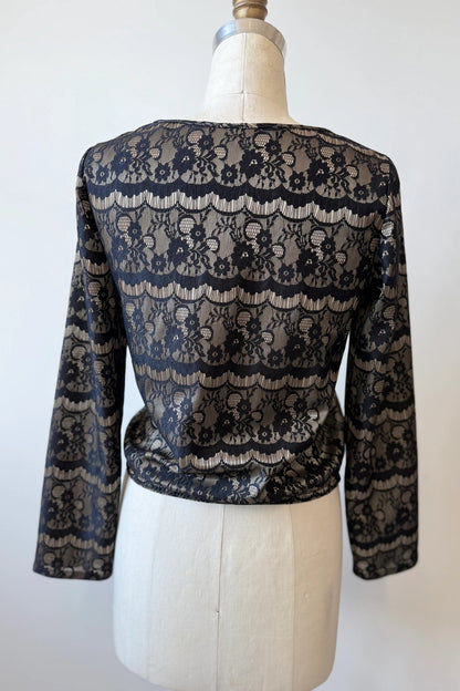 A back view of the Solstice Blouse by Kollontai in Gold is shown on a mannequin in front of a white background.  The fabric layers black lace over golden satin. It's slightly cropped with an elastic waist and long sleeves. 
