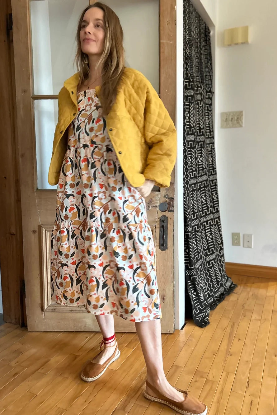 Woman wearing the Artemesia Jacket by Kazak in Mustard, a quilted jacket with snaps up the front and large patch pockets. She is wearing it over  the Lavendula Dress in Cream with Floral and standing in a room with wooden floor door. 