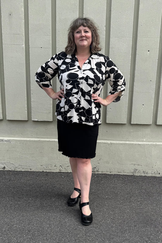 A woman wearing the Francesca Blouse by Pure in Black/Ivory geometric print, an oversized blouse with a lapel collar, button front, 3/4 sleeves with button and tab detail, and rounded h-low hemline. She is wearing it with a black skirt and standing against a grey wall.