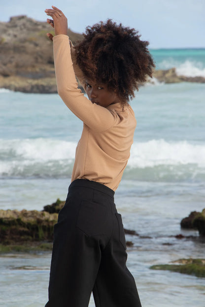 Side view of a woman wearing the Vienna Pants by MAS in Pepper, wide legged pants with a high waist and a zip front. She is wearing them with a beige top and standing on a rocks by the water. 