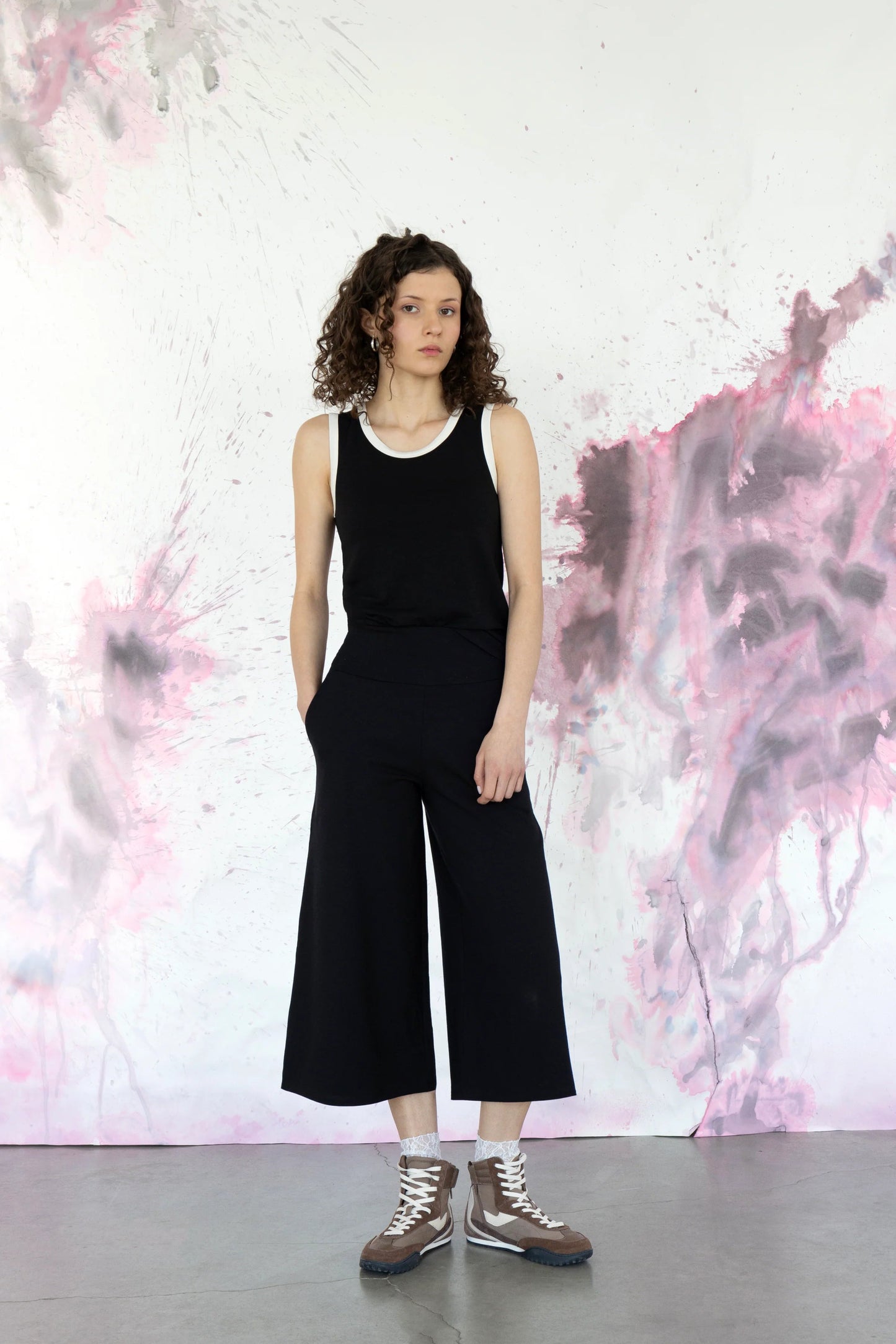A woman wearing the Trille Trousers by Eve lavoie in Burgundy, wide legged 3/4 length pants with a wide pull-on waistband. She is wearing them with a black tank and standing in front of a speckled purple background. 