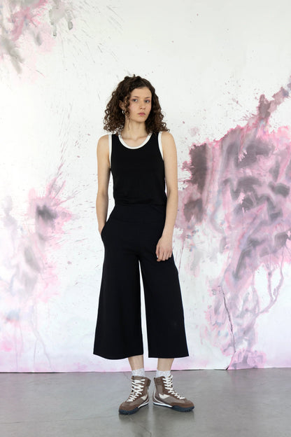 A woman wearing the Pissenlit Camisole by Eve Lavoie in Black/White, a black tank with white contrast piping at the neck and shoulders. She is wearing it with the Trille Trousers in Black and standing against a abstract pink and grey wall.