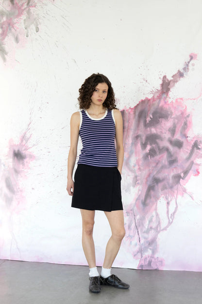 A woman wearing the Pissenlit Camisole by Eve Lavoie in Lavender Stripe, a  striped tank top with white piping at the neck and shoulders.  She is wearing it with the Campanule Skort in Black and standing against a abstract pink and grey wall. 