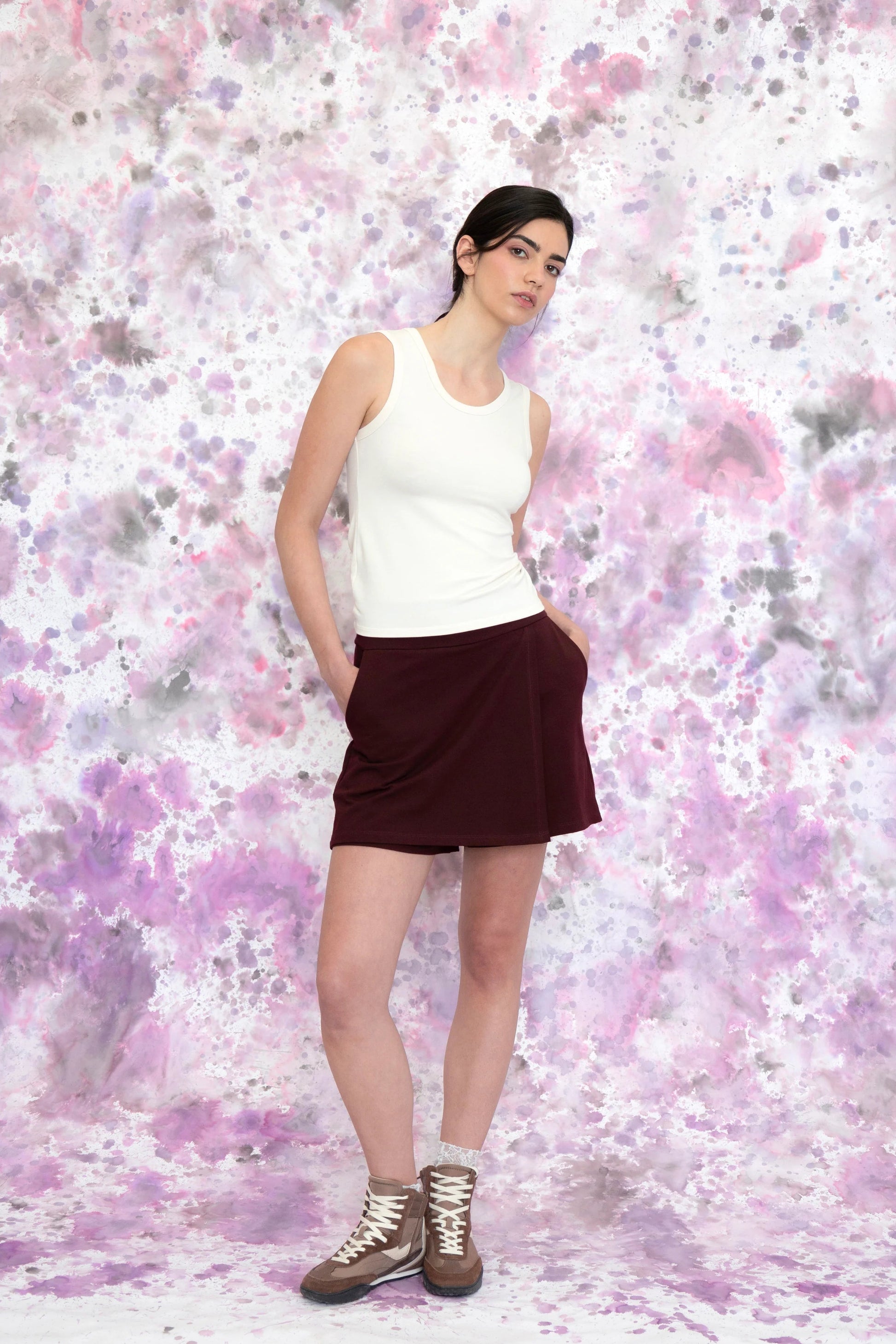 A woman wearing the Campanule Skort by Eve Lavoie in Burgundy, a short pull on skirt with a faux-wrap skirt effect in the front. She is wearing it with a white tank and standing in front of a speckled purple and white background.