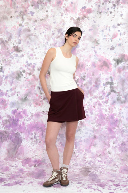 A woman wearing the Campanule Skort by Eve Lavoie in Burgundy, a short pull on skirt with a faux-wrap skirt effect in the front. She is wearing it with a white tank and standing in front of a speckled purple and white background.
