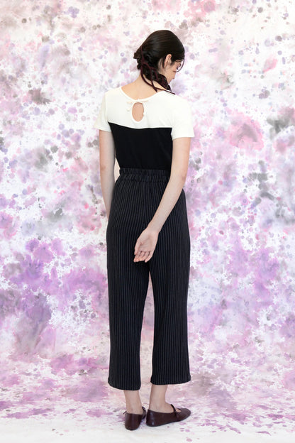 Back view of a woman wearing the Tanaisie Woven Pants from Eve Lavoie in Black Stripe, slightly cropped linen/cotton pants with wide legs, an elastic waist, classic fly closure, and slanted side pockets. She is wearing them with a black and white top and standing in front of a purple and white speckle background. 