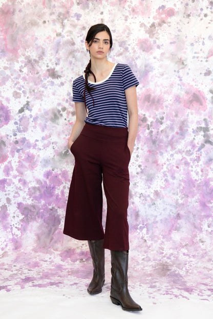 A woman wearing the Monarde Top by Eve Lavoie in Lavender Stripe with white piping. It is reversible back to front, with U-neckline on one side and a boat neckline on the other. She is wearing it with the Trille Trousers in  Burgundy pants and standing against an abstract background