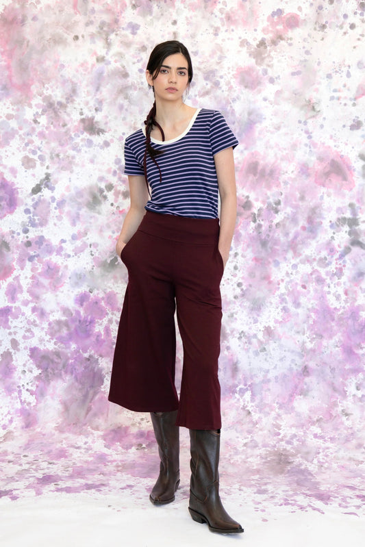 A woman wearing the Monarde Top by Eve Lavoie in Lavender Stripe with white piping. It is reversible back to front, with U-neckline on one side and a boat neckline on the other. She is wearing it with the Trille Trousers in  Burgundy pants and standing against an abstract background
