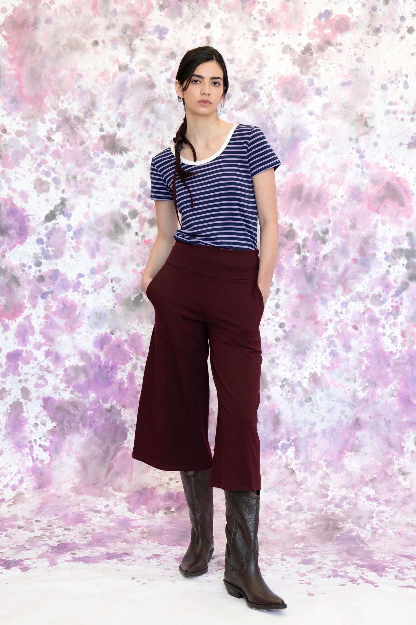 A woman wearing the Monarde Top by Eve Lavoie in Lavender Stripe with white piping. It is reversible back to front, with U-neckline on one side and a boat neckline on the other. She is wearing it with the Trille Trousers in  Burgundy pants and standing against an abstract background