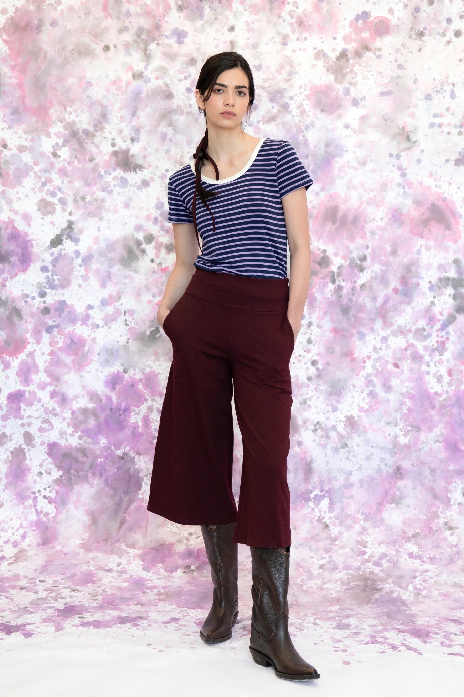 A woman wearing the Monarde Top by Eve Lavoie in Lavender Stripe with white piping. It is reversible back to front, with U-neckline on one side and a boat neckline on the other. She is wearing it with the Trille Trousers in  Burgundy pants and standing against an abstract background