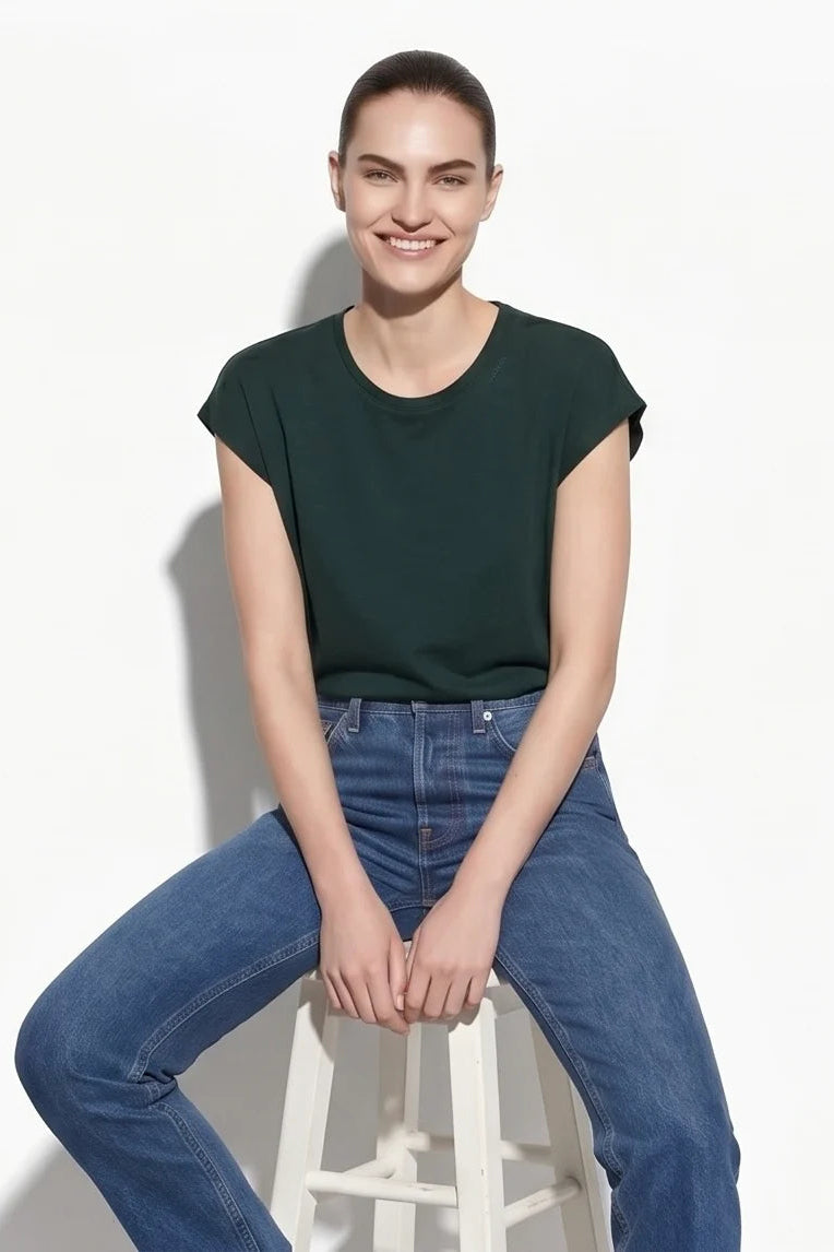 A woman wearing the Eowyn T-Shirt by Kollontai in Green with jeans sitting on a stool against a grey background. 