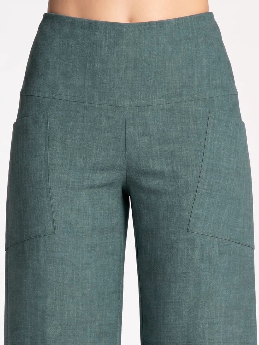 Close up waist down view of a woman wearing the Kollontai Susan Pants in Green, standing in front of a grey background. 