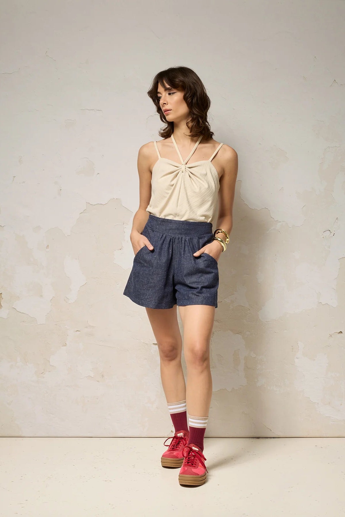 A woman wearing the Maya shorts from Cokluch in Indigo Speckle, lightweight denim shorts with a waist that's elasticized at the back, wide culotte style legs, and a side pockets. She is wearing them with a white tank and standing in front of a grey background. 