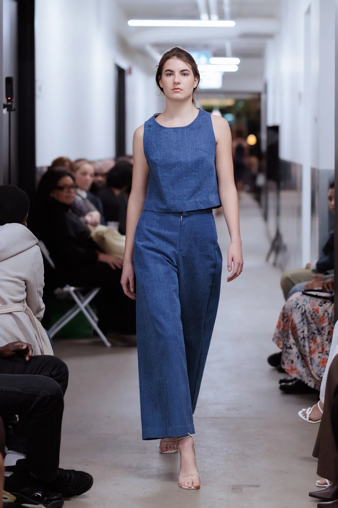 A woman wearing the Vienna Pants by MAS in Denim,  wide legged pants with a high waist and a zip front. She is wearing them with a matching top and  walking on a catwalk with an audience seated on either side.