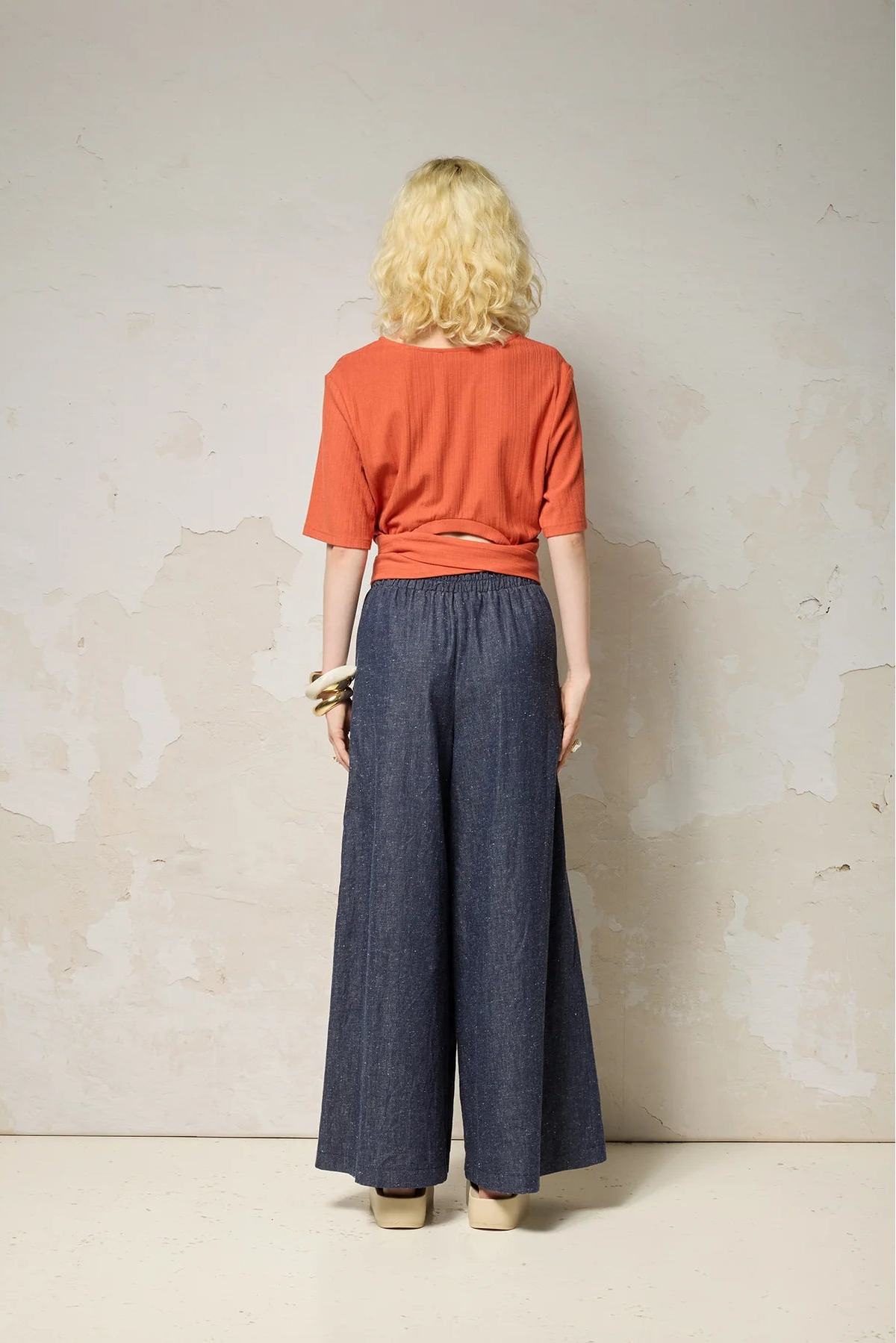 Back view of a woman wearing the Kali Top by Cokluch in Fiesta, a ribbed knit top with elbow-length sleeves, an integrated built, and an opening at the lower back. She is wearing it with blue pants and standing against a neutral background