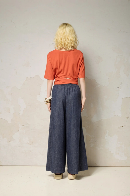 Back view of a woman wearing the Kali Top by Cokluch in Fiesta, a ribbed knit top with elbow-length sleeves, an integrated built, and an opening at the lower back. She is wearing it with blue pants and standing against a neutral background