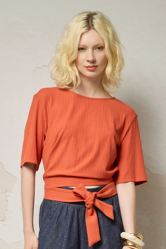 Waist up view of a woman wearing the Kali Top by Cokluch in Fiesta, a ribbed knit top with elbow-length sleeves, an integrated built, and an opening at the lower back. She is wearing it with blue pants and standing against a neutral background.