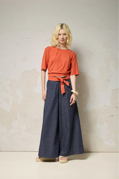 A woman wearing the Kali Top by Cokluch in Fiesta, a ribbed knit top with elbow-length sleeves, an integrated built, and an opening at the lower back. She is wearing it with blue pants and standing against a neutral background