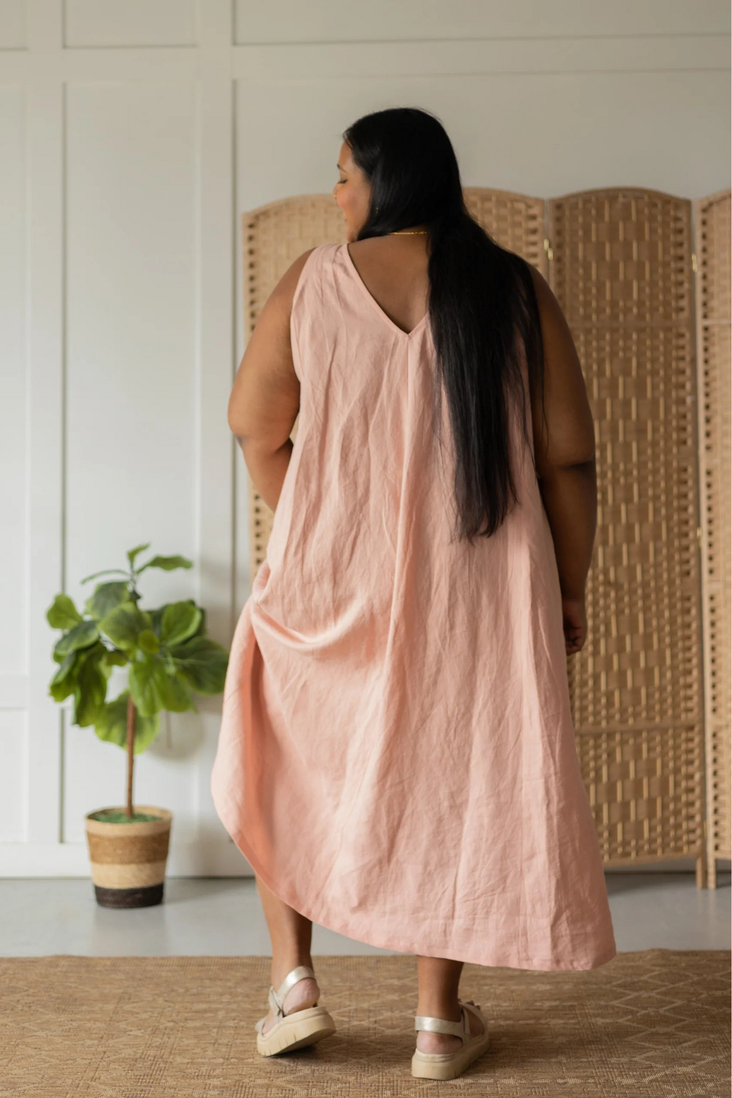  Back view of a woman wearing the Caroline Dress by Lights of All in Ballet Pink, a sleeveless dress linen maxi dress with a V-neck at the back. She is standing against a white wall with a woven screen behind her. 