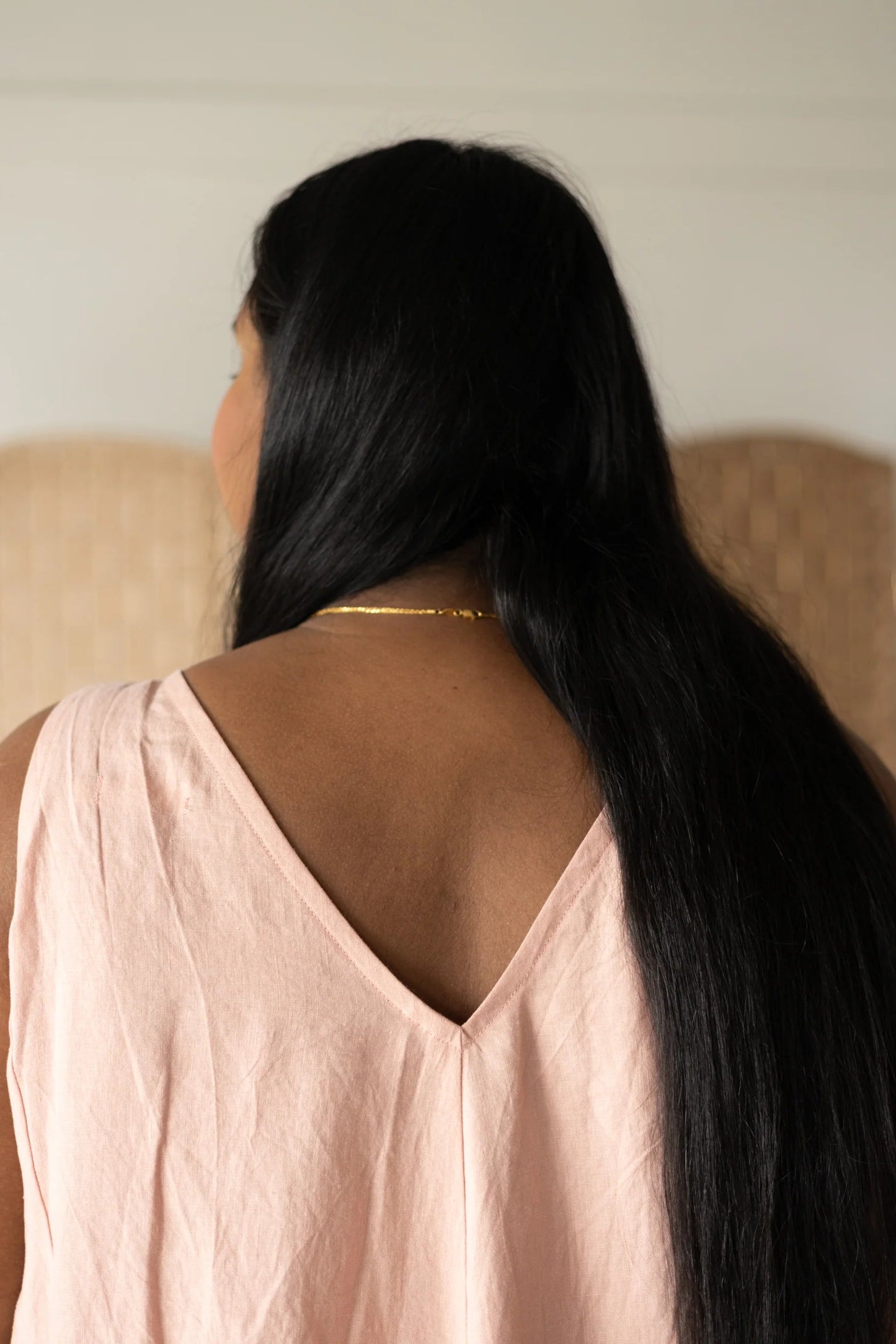 Back close up view of a woman wearing the Caroline Dress by Lights of All in Ballet Pink, a sleeveless dress linen maxi dress with a V-neck at the back. She is standing against a white wall with a woven screen behind her. 