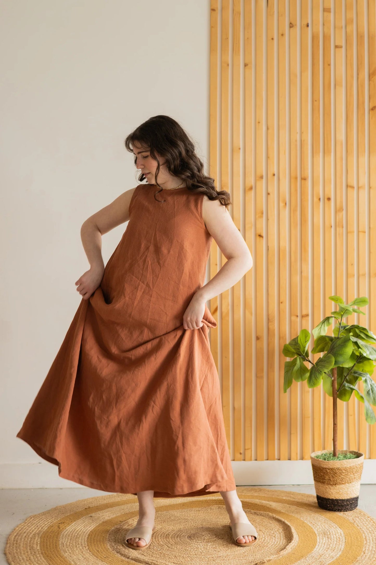 A woman wearing the Caroline Dress by Lights of All in Sedona, a sleeveless dress linen maxi dress with a V-neck at the back. She is standing against a white and wood panelled wall. 