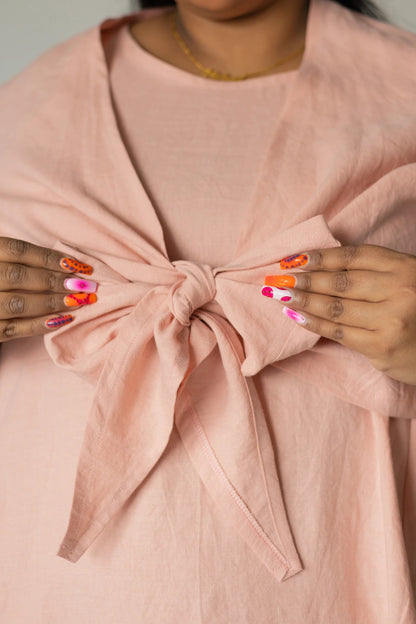 A woman wearing the Stella Top by Lights of All in Ballet Pink, a short sleeved linen wrap top that can be twisted or tied back or front. She is wearing it over the matching Caroline dress and holding the bow.