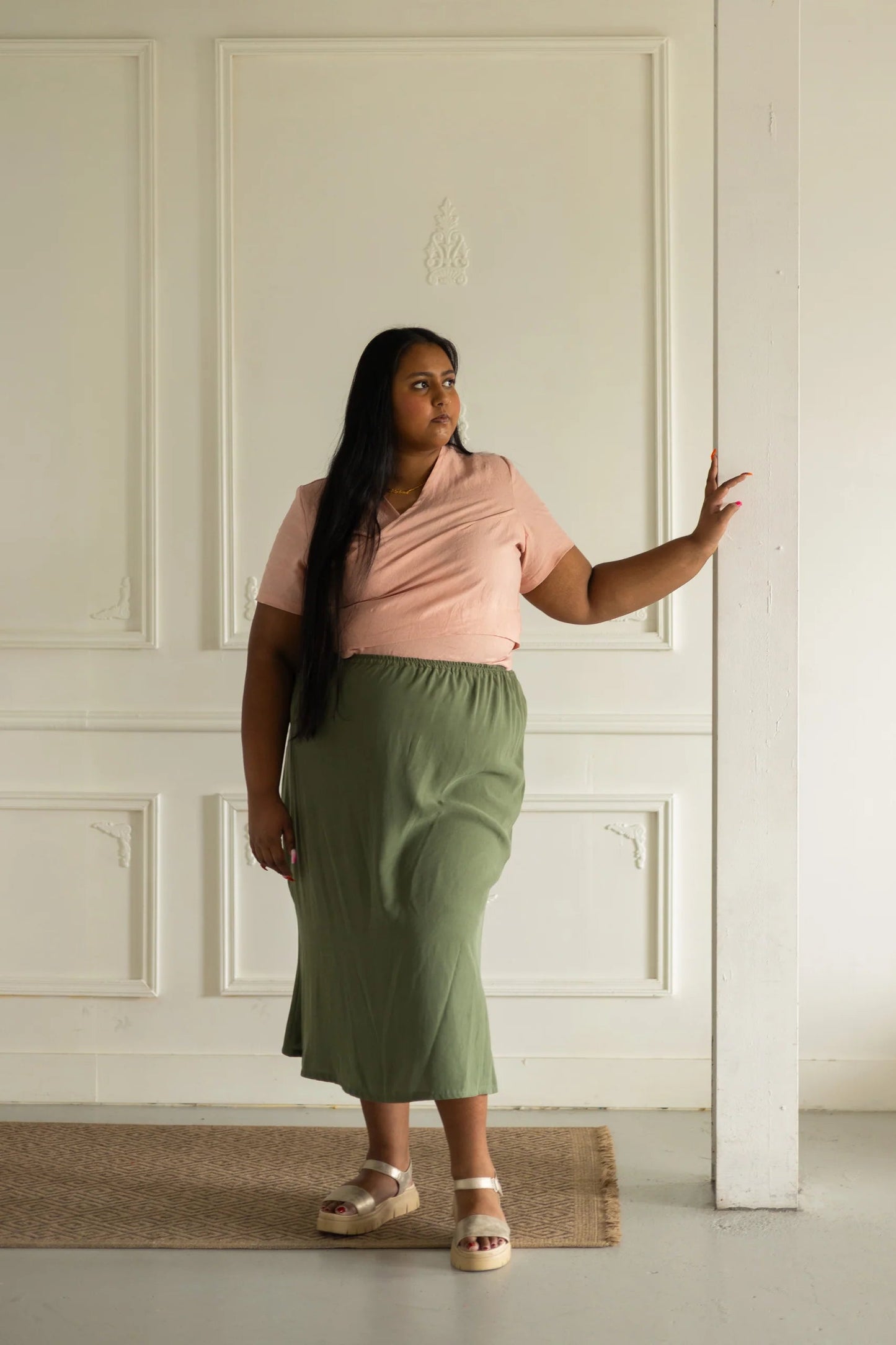 A woman wearing the Simone Skirt by Lights of All in Cactus, a sleek calf-length skirt with a flared hem and an elastic waist. She is wearing it with the Stella Top in Ballet Pink, and standing in a white room.