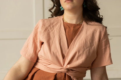 Torso view of a woman wearing the Stella Top by Lights of All in Ballet Pink, a short sleeved linen wrap top that can be twisted or tied back or front. She is wearing it over the Caroline dress in Sedona and sitting in a white room. 
