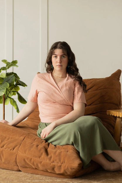 A woman wearing the Simone Skirt by Lights of All in Cactus, a sleek calf-length skirt with a flared hem and an elastic waist. She is wearing it with the Stella Top in Ballet Pink and sitting on a brown chair in a white room. 