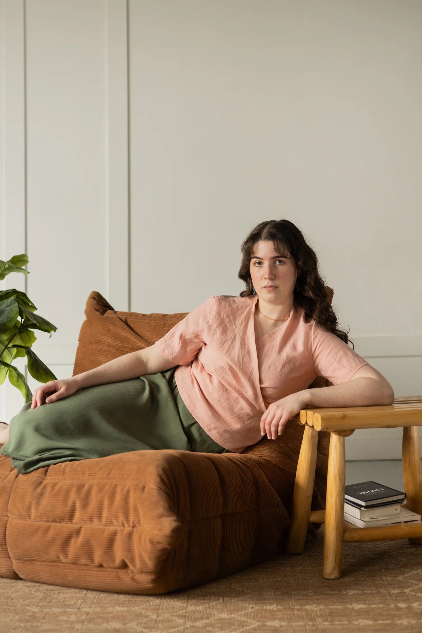 A woman wearing the Stella Top by Lights of All in Ballet Pink, a short sleeved linen wrap top that can be twisted or tied back or front. She is wearing it over the Simone Skirt in Cactus Sedona and sitting on a brown chair in a white room. 