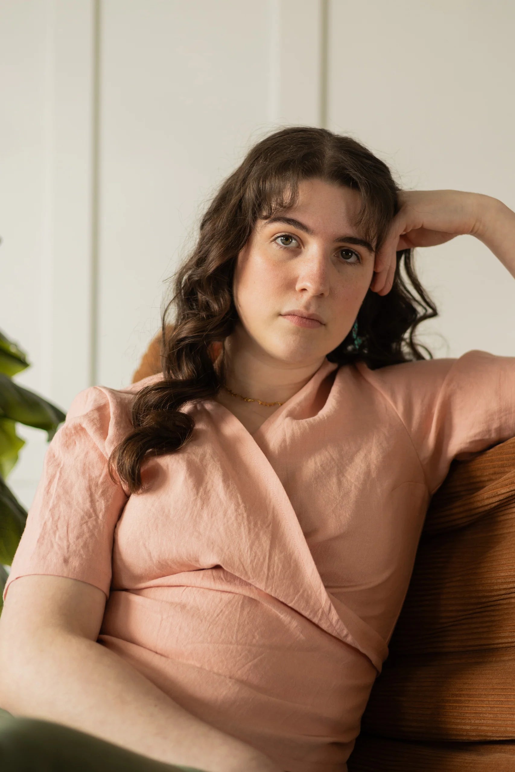 A woman wearing the Stella Top by Lights of All in Ballet Pink, a short sleeved linen wrap top that can be twisted or tied back or front. She is wearing it over the Simone Skirt in Cactus Sedona and sitting on a brown chair in a white room. 