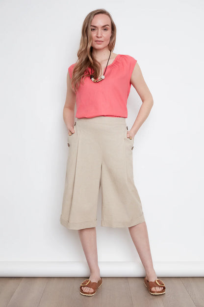 A woman wearing the Isadora Capri Pants by Julei in Natural, wide-leg cropped pants with three coconut buttons along each pocket. She's wearing them with a coral sleeveless top and standing in front of a white background. 