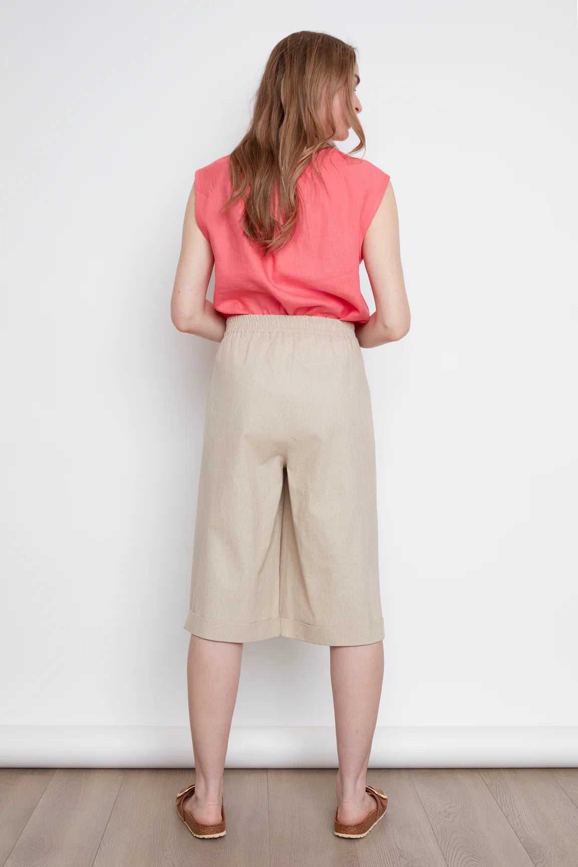 Back view of a woman wearing the Isadora Capri Pants by Julei in Natural, wide-leg cropped pants with three coconut buttons along each pocket. She's wearing them with a coral sleeveless top and standing in front of a white background. 