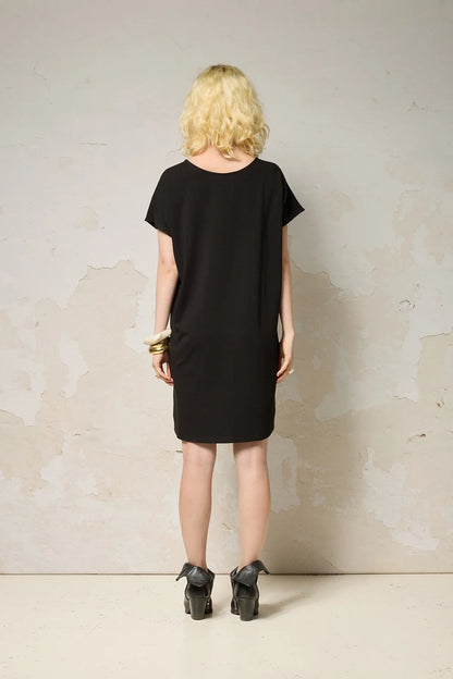 Back view of a woman wearing the Lyssa Dress by Cokluch in Black, a short dress with a fringed v-panel at the cheset, short sleeves, and a relaxed fit. She is standing against a textured wall.