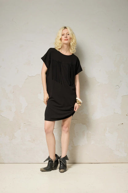 A woman wearing the Lyssa Dress by Cokluch in Black, a short dress with a fringed v-panel at the cheset, short sleeves, and a relaxed fit. She is standing against a textured wall.