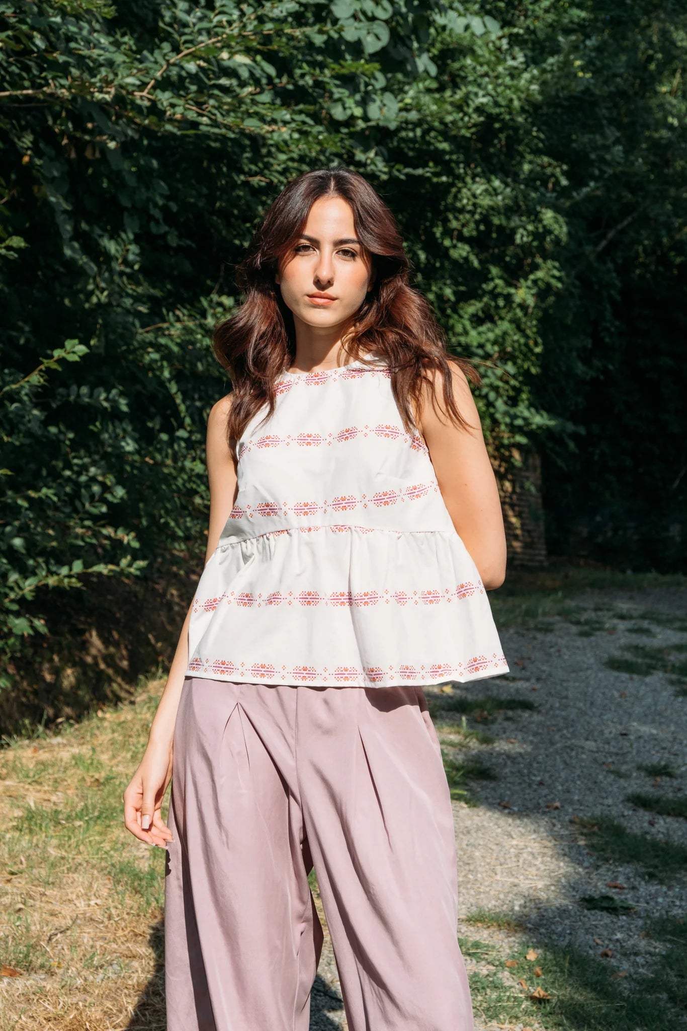 Woman wearing the Malé Top from MAS, a sleeveless top with a peplum and ties up the back. She is wearing it with wide-leg pants standing outdoors with greenery in the background. 