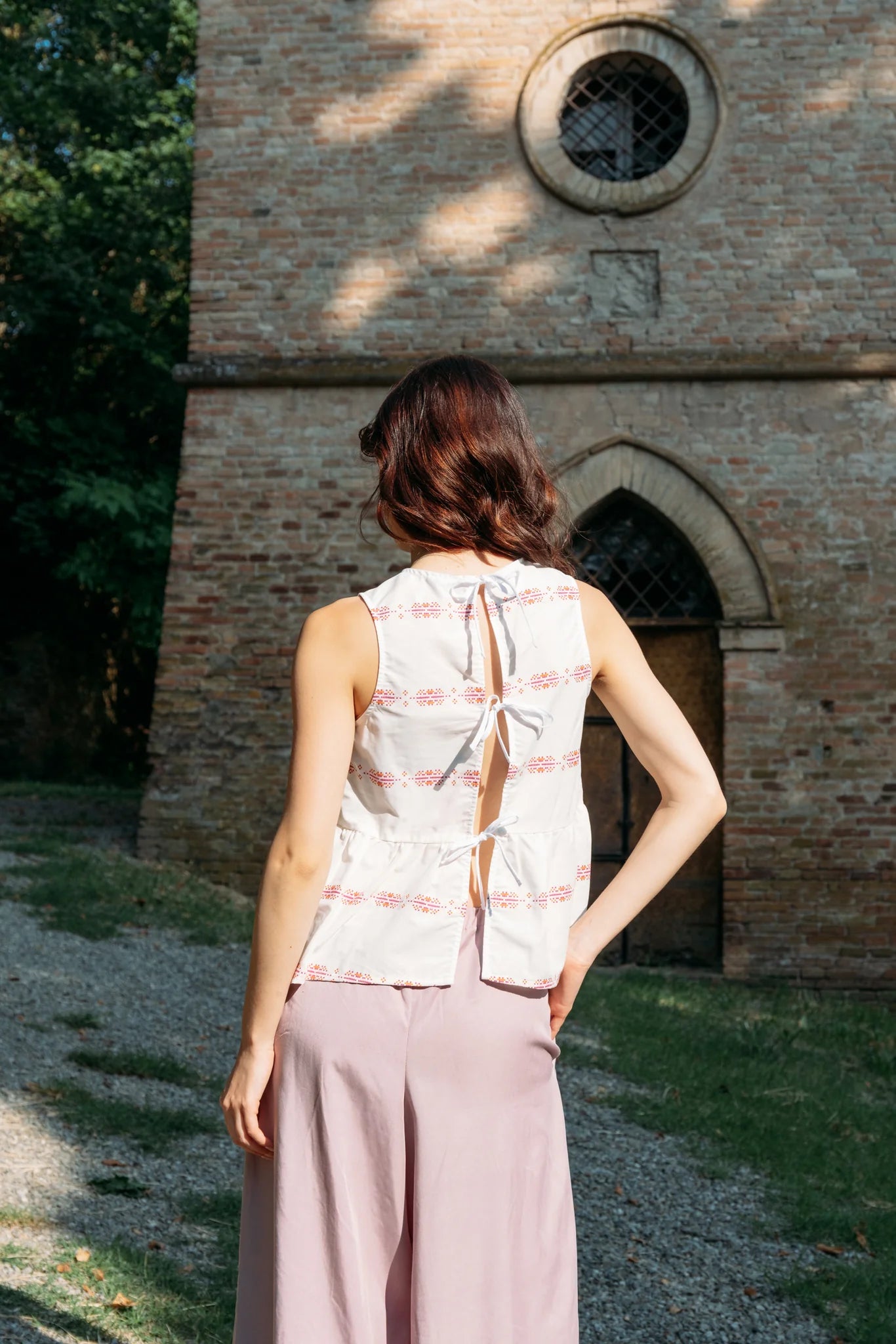 Back view of a woman wearing the Malé Top from MAS, a sleeveless top with a peplum and ties up the back. She is wearing it with wide-leg pants standing outdoors with a stone building in the background. 