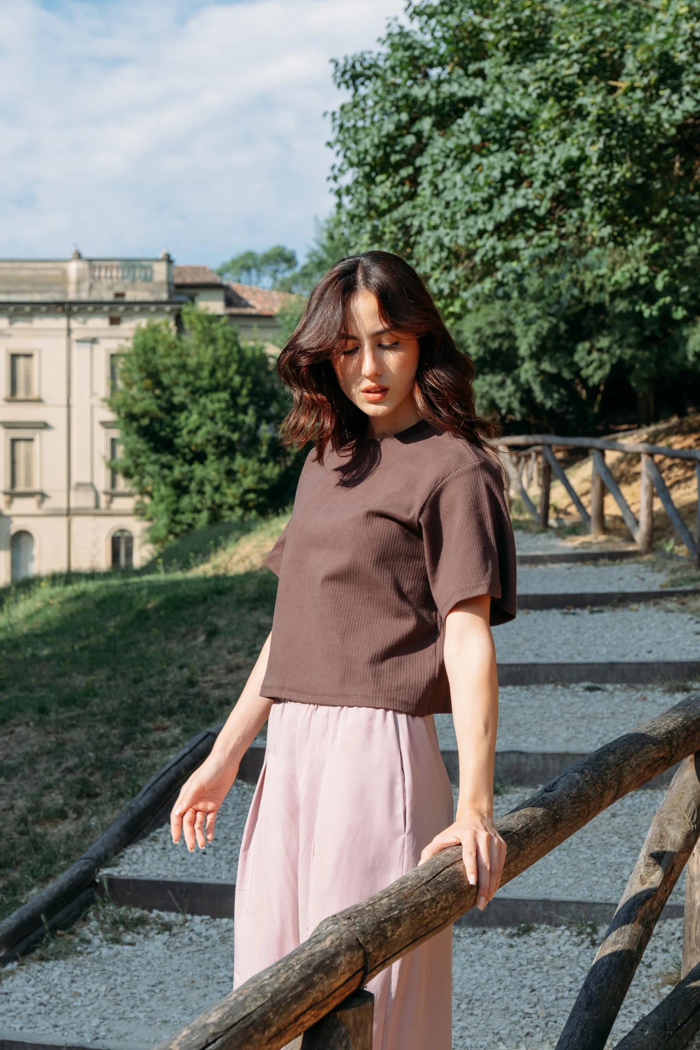 A woman wearing the Siena Pants by MAS in Raspberry, wide legged trousers with an elastic waist. She is wearing them with a brown top and standing outdoors on a fenced pathway.
