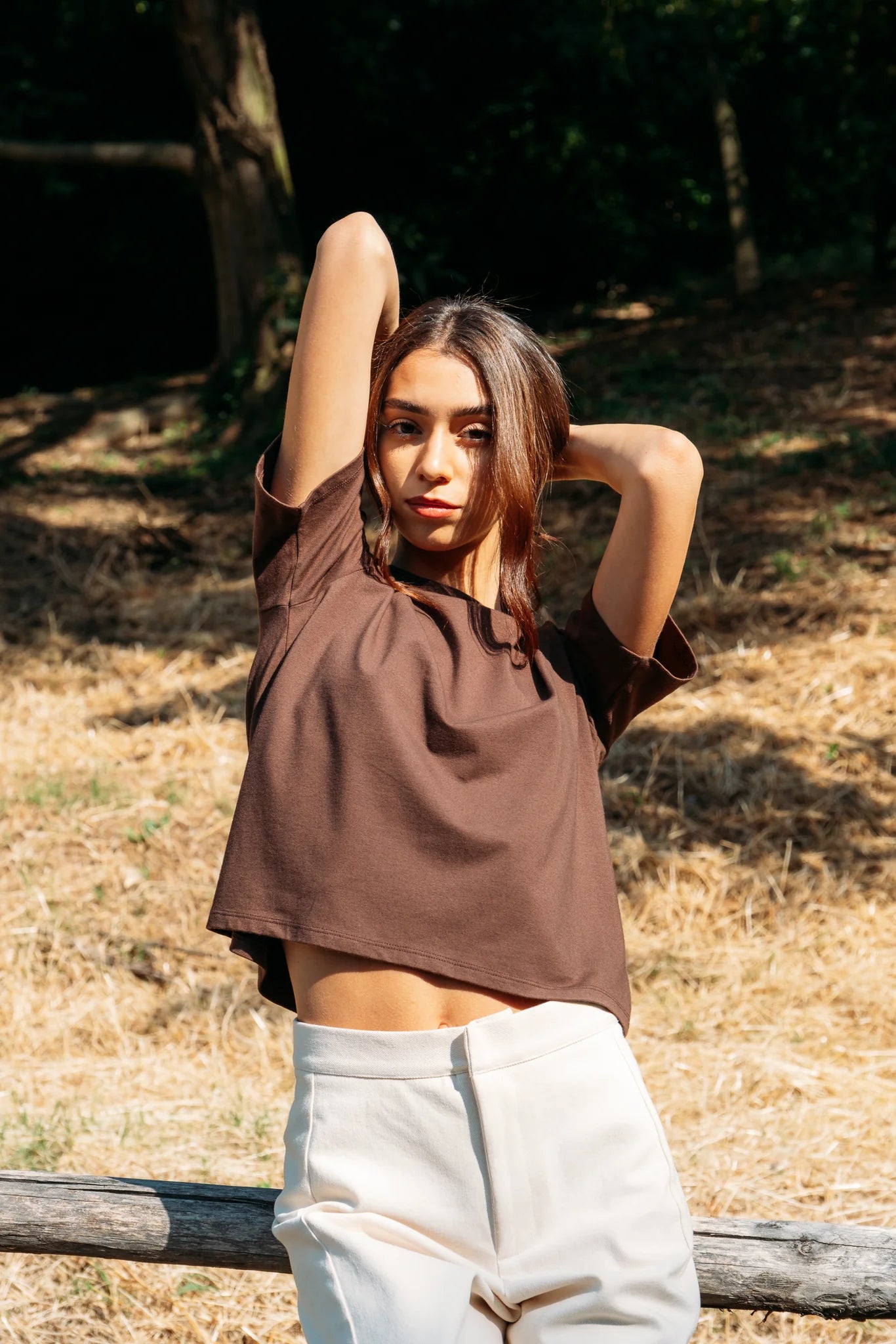 A woman wearing the Santiago T-Shirt by MAS in Cacao, a classic tee with a slightly cropped and boxy fit. She is wearing it with beige pants leaning on a log fence with a forest behind her. 