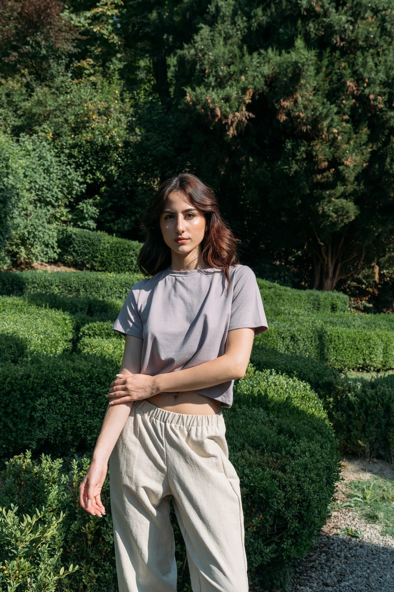 A woman wearing the Santiago T-Shirt by MAS in Lilac, a classic tee with a slightly cropped and boxy fit. She is wearing it with beige pants and standing outside with greenery behind her. 
