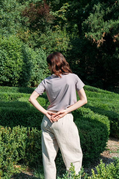 Back view of a woman wearing the Santiago T-Shirt by MAS in Lilac, a classic tee with a slightly cropped and boxy fit. She is wearing it with beige pants and standing outside with greenery behind her. 