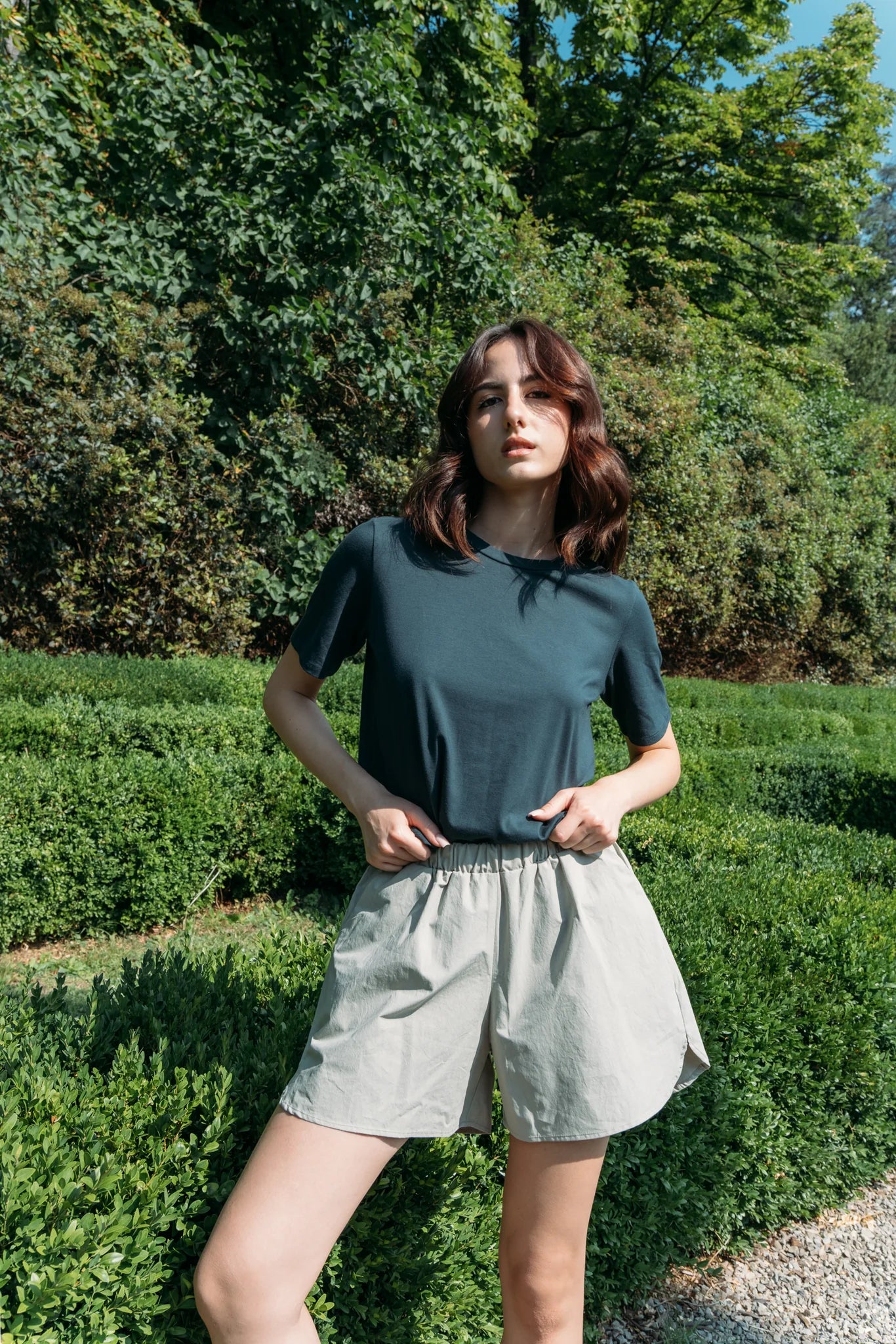 A woman wearing the Santiago T-Shirt by MAS in Emerald, a classic tee witha slightly cropped and boxy fit. She is wearing it with beige shorts and standing outside with greenery behind her. 