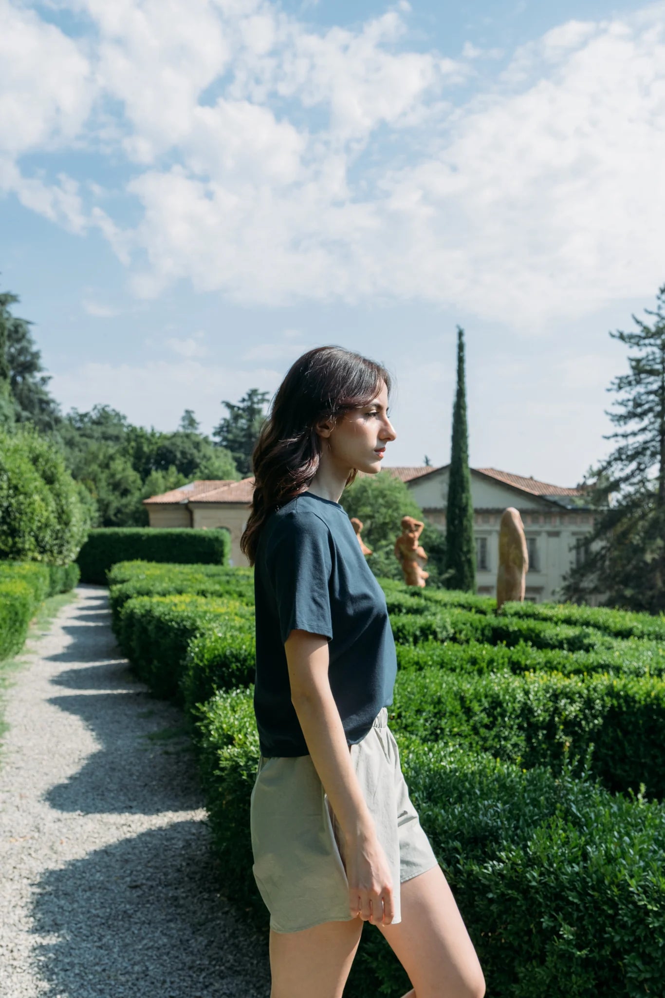 Side view of a woman wearing the Santiago T-Shirt by MAS in Emerald, a classic tee witha slightly cropped and boxy fit. She is wearing it with beige shorts and standing outside with greenery behind her. 