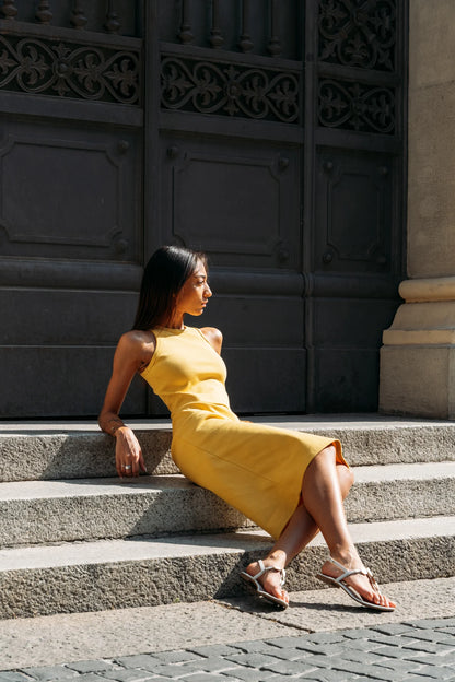 A woman in wearing the Lima Tank by MAS in Lemon, a cropped rib tank with a high neck and wide straps. She is wearing it with a matching skirt and sitting on stone steps outdoors.