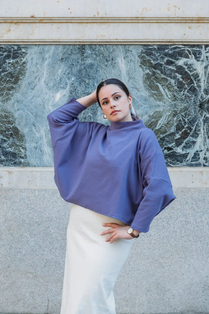 Woman wearing the Bogota Top by MAS in Violet, an oversized top with a high neck, exaggerated dropped shoulders, and long sleeves, in ribbed fabric. She is wearing it with a white skirt and standing in front of a white wall with a cracked window.  