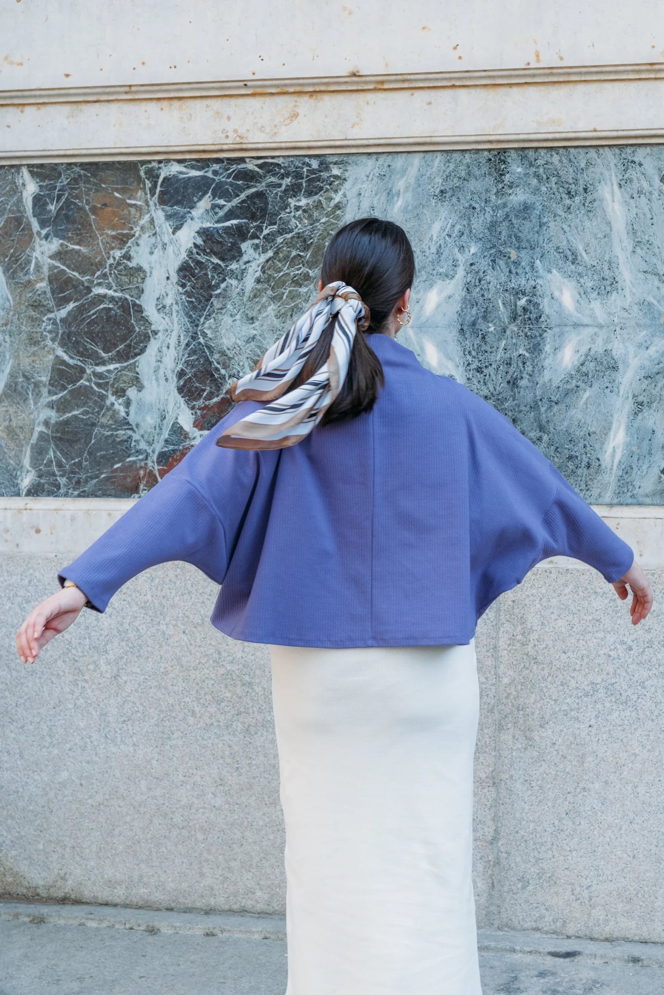 Back view of a woman wearing the Bogota Top by MAS in Violet, an oversized top with a high neck, exaggerated dropped shoulders, and long sleeves, in ribbed fabric. She is wearing it with a white skirt and standing in front of a white wall with a cracked window.  