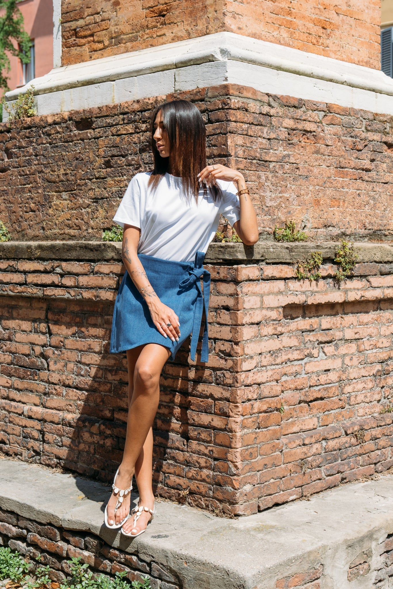 A woman wearing the Santiago T-Shirt by MAS in Salt, a classic tee with a slightly cropped and boxy fit. She is wearing it with a denim skirt and standing outside leaning against a. brick building. 